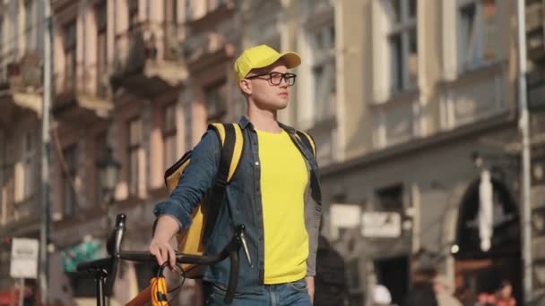 Een jonge gelukkige bezorger gaat en houdt een fiets bij zich. De wind blaast zijn shirt op. Hij draagt gele kleding. Hij draagt een gele rugzak voor levering op zijn schouders. 4K — Stockvideo