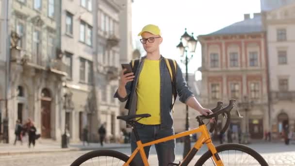 Portret van een vrolijke bezorger die staat en sms 't op een smartphone. Hij steekt zijn hoofd op en kijkt naar de camera. Een fiets vasthouden. Hij draagt een gele rugzak op zijn schouders. 4K — Stockvideo
