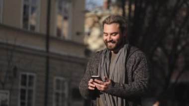 Mutlu sakallı bir adam akıllı telefondan sosyal ağlara göz atıyor. Süveter ve eşarp giyiyor. O, eski kentin merkezinde duruyor. Güneş parlıyor. 4K