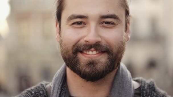 Портрет бородатого счастливого человека, смотрящего в камеру и улыбающегося. Его волосы качаются на ветру. Старый центр города на заднем плане. 4K — стоковое видео