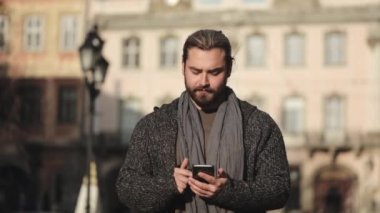 Sakallı bir adam akıllı telefondan sosyal ağlara girip başını kaldırıyor. Gülümsüyor. Süveter ve eşarp giyiyor. O, eski kentin merkezinde duruyor. 4K