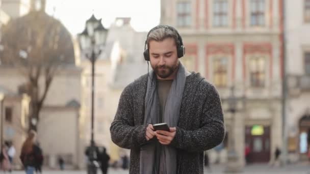 一个快乐的大胡子男人正在用耳机听音乐，并在智能手机上浏览社交网络。他面带微笑,穿过市中心.4K — 图库视频影像