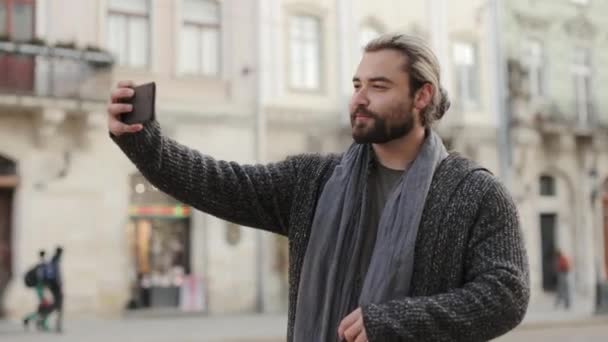 Um homem barbudo feliz está tirando uma selfie no centro da cidade velha e ele aponta um sinal de paz com os dedos. Ele está a sorrir. Está a usar uma camisola. O sol está brilhando intensamente. 4K — Vídeo de Stock