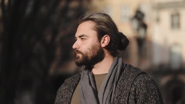 Портрет бородатого человека, который поворачивает голову и смотрит в камеру. Он носит свитер и шарф. Он стоит в центре старого города. 4k — стоковое видео