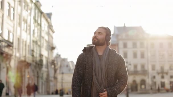 Um homem barbudo feliz está ouvindo música e andando pelo centro da cidade. Ele está sorrindo e olhando ao redor. O sol está brilhando intensamente. Está a usar uma camisola. 4K — Vídeo de Stock