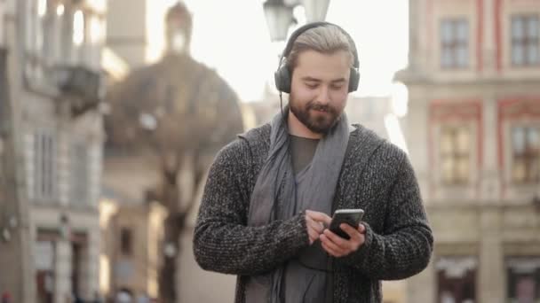 A happy bearded man is listening to music with headphones and scrolling through social networks on his smartphone. He is smiling and raising his head. The old town is in the background. 4K — Video