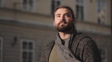 Sakallı bir adam şehir merkezinde yürüyor. Gülümsüyor ve etrafa bakıyor. Güneş ışıl ışıl parlıyor ve rüzgar esiyor. 4K