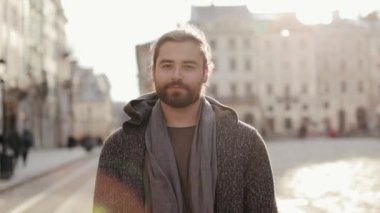 Sakallı bir adamın portresi kameraya bakıyor ve gülümsüyor. O, eski şehir merkezinde duruyor. Süveter ve eşarp giyiyor. Güneş ışıl ışıl parlıyor. 4K