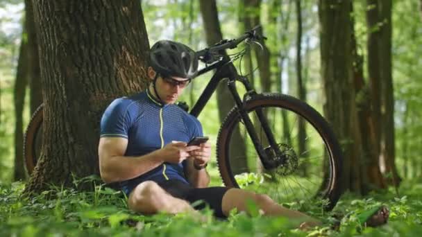 Un ciclista è seduto vicino a un albero e sta navigando sui social network. Sta riposando dopo un allenamento. Indossa vestiti da ciclista. MTB bike è in piedi sullo sfondo. 4K — Video Stock