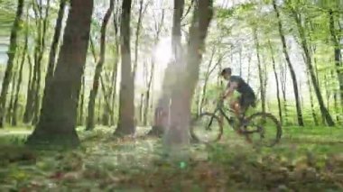 MTB motorlu bir bisikletçi yüksek hızda ormanda ilerliyor. Kamera ona paralel hareket ediyor. Güneş ağaçların arasından parlıyor. 4K