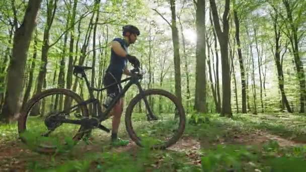 Un hombre feliz con una bicicleta está caminando a lo largo de un sendero forestal. Lleva un uniforme de ciclista. El sol brilla intensamente. Entrenamiento en una bicicleta MTB. 4K — Vídeo de stock