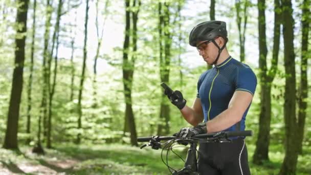 Un uomo sta guardando attraverso i social network su uno smartphone. E 'in piedi accanto a una bici MTB nel bosco. Indossa vestiti da ciclista. La telecamera si muove da sinistra a destra. 4K — Video Stock