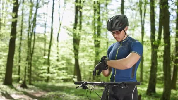 サイクリストは時計のトレーニングデータを見ています。森の中のMTBバイクの隣に立っている。彼はサイクリング服を着ている。カメラは左から右に動いている。4K — ストック動画