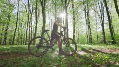 Bisikletli bir bisikletçi orman yolunda yürüyor. Bisiklet üniforması giyiyor. Güneş ağaçların arasından ışıl ışıl parlıyor. MTB bisiklet eğitimi. 4K