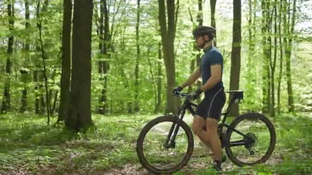 Ciclistul se plimbă lângă bicicletă. Se uită în jurul pârtiei forestiere. Poartă echipament de ciclism. Camera se mişcă cu el. 4K — Videoclip de stoc
