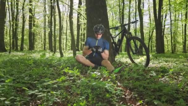 サイクリストはスマートフォンでソーシャルネットワークを見ています。彼は木のそばに座っている。彼の隣に自転車がある。サイクリングトレーニングの後に休憩。太陽は明るく輝いている。4K — ストック動画