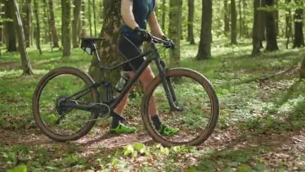 Un ciclista está caminando con una bicicleta a lo largo de un sendero forestal. Lleva ropa de ciclista. La cámara está enfocada en la moto. 4K — Vídeos de Stock