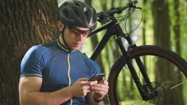 Un ciclista está viendo análisis de entrenamiento en un teléfono inteligente. Está sentado junto a un árbol. Hay una bicicleta a su lado. El sol brilla intensamente. 4K — Vídeo de stock