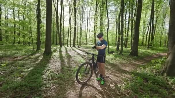 Szczęśliwy rowerzysta stoi w środku lasu i patrzy na dane treningowe ze swojej wachty. Obok niego jest rower. Ma na sobie mundur rowerowy. 4K 50fps — Wideo stockowe