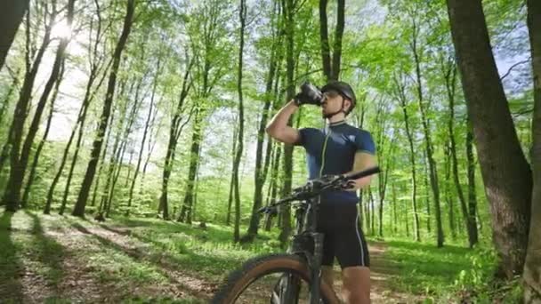 Чоловік на велосипеді стоїть на лісовій стежці. Він п'є воду з пляшки. Він відпочиває після тренування. Камера рухається навколо нього. 4k — стокове відео