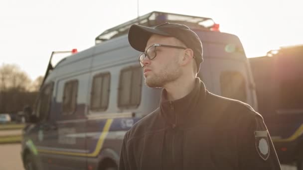 Um homem de uniforme policial está parado perto de dois ônibus da polícia e inspecionando a área. Está a usar óculos. O sol está brilhando intensamente. 4K — Vídeo de Stock