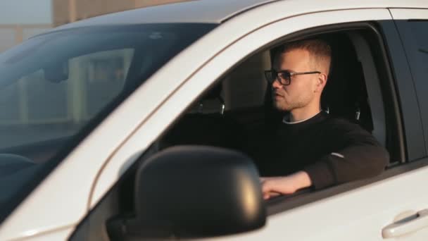 Um homem feliz está sentado em um carro e segurando a mão na porta. Ele está virando a cabeça e olhando para a câmera. Está a usar óculos. 4K — Vídeo de Stock