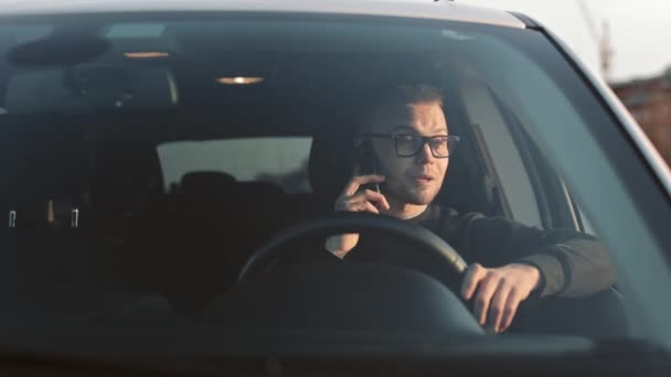 Un hombre está sentado en un coche y hablando emocionalmente por teléfono. Mantiene su mano en el volante. Lleva gafas puestas. Disparando por el parabrisas. 4K 50fps — Vídeo de stock