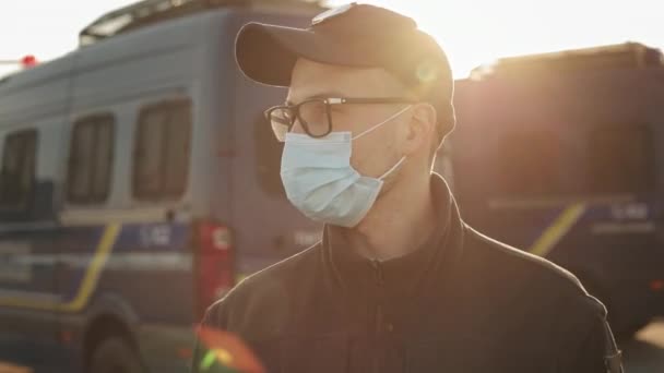 Un homme en uniforme de police se tient près de deux bus de police. Il porte un masque de protection. Il portait des lunettes. Le soleil brille de mille feux. 4K — Video