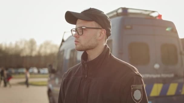 Muž v policejní uniformě stojí u dvou policejních autobusů a rozhlíží se. Má na sobě brýle. Slunce jasně svítí. 4K — Stock video