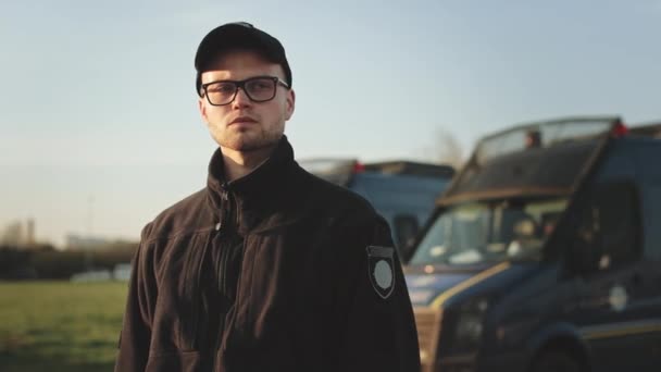 Muž v policejní uniformě stojí a rozhlíží se. Má na sobě brýle. V pozadí jsou dva policejní autobusy. 4K — Stock video