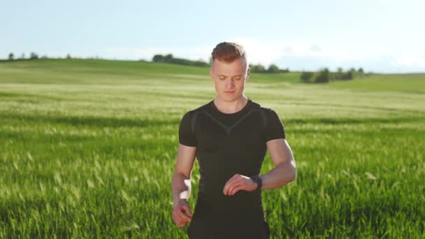 Um jovem de uniforme desportivo está em campo. Ele está olhando para as estatísticas de treinamento no relógio de fitness. Ele está levantando a cabeça e desviando o olhar. Tiro de retrato. 4K — Vídeo de Stock