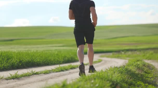 Um jovem atleta corre rapidamente ao longo de uma estrada de campanha. Ele está a treinar para se manter em forma. Campos com grama verde no fundo. A câmara está a filmá-lo por trás. 4K — Vídeo de Stock