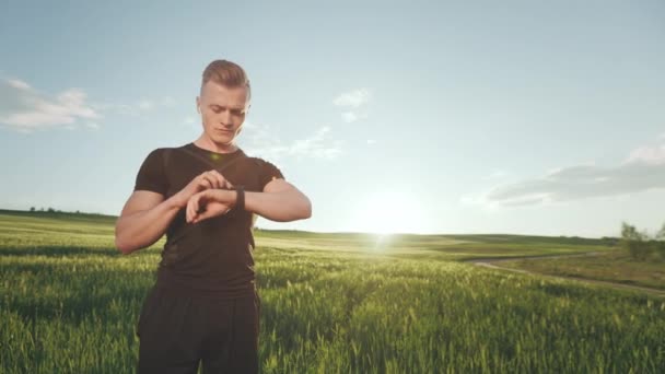 Een jongeman in een sportuniform staat op het veld. Hij bekijkt de trainingsstatistieken op het fitnesshorloge. Portret schieten. 4K — Stockvideo