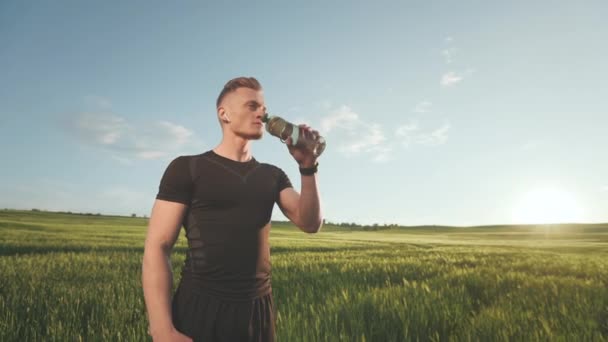 Спортсмен стоит в поле и пьет воду из бутылки. Он носит спортивную форму. Закат. Портретная съемка. 4K — стоковое видео