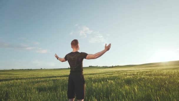 Um homem de uniforme desportivo está no meio de um campo. Ele está olhando alegremente para o pôr do sol e segurando as mãos no ar. A câmara está a dar-lhe a volta. 4K — Vídeo de Stock