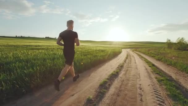 Un sportif court le long d'un chemin de terre près d'un champ. Il portait un uniforme de sport. Coucher de soleil. 4K — Video