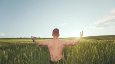 Tişörtsüz mutlu bir adam bir tarlanın ortasında oturuyor. Gün batımına bakıyor ve ellerini havada tutuyor. Doğanın tadını çıkarıyor. 4K