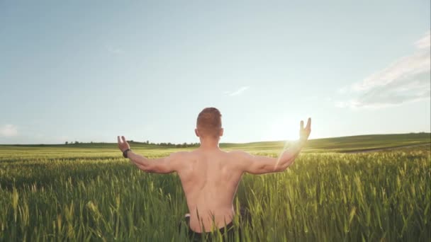 Un homme heureux sans t-shirt est assis au milieu d'un champ. Il regarde le coucher du soleil et tient les mains en l'air. Il apprécie la nature. 4K — Video