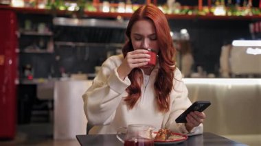 Beyaz giyinmiş, sıcak içkisini yudumlayan, gözleri kapalı, huzurlu bir kadın. Akıllı telefonu elinde tutarak. Sessiz kafe ziyareti sırasında ara vermenin keyfini çıkarırken haber okumak veya kayan sosyal medya.