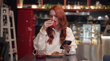 Güzel, beyaz bir kadın akıllı telefondayken sıcak içkinin tadını çıkarıyor. Önünde tatlıyla kafe masasında oturmak. Yavaşça yudumluyor. Metin okunuyor ya da iletiye sakin bir ifadeyle yanıt veriliyor.