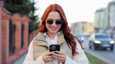 Kızıl saçlı, açık havada duran ve akıllı telefonuyla fotoğraf çeken beyaz bir kadın. Gülümseyen bir kadın, arka planda binalar ve arabalarla şehir caddesinde anı yakalıyor. Gülümsüyor.