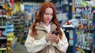 Gülümseyen beyaz kadın mağazada duruyor ve akıllı telefon kullanıyor. Kırmızı saçlı, bej yelekli bir kadın evin içinde renkli paketlerle dolu raflarla alışveriş yaparken mesaj yazıyor..