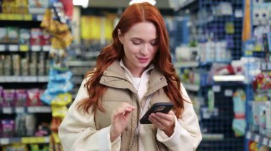 Mağazanın içinde akıllı telefon kullanan bej renkli yelekli kızıl saçlı beyaz kadın. Gülümseyen kadın cep telefonundan ürün bilgilerini kontrol ediyor. Etrafı renkli mallarla çevrili raflarla çevrili alışveriş..