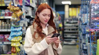 Mağaza koridorunda dikilirken kızıl saçlı, akıllı telefonlu sakin bir beyaz kadın. Bej yelek denetim listesindeki kadın veya raflardaki renkli ambalajlarla çevrili ürünleri karşılaştırma.