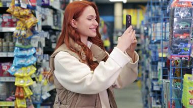Mağaza koridorunda durup akıllı telefondan fotoğraf çeken kızıl saçlı, gülümseyen beyaz kadın. Bej renkli yelek giyen kadın, kapalı alanda renkli ambalajlarla çevrili ürünün görüntüsünü yakalıyor..