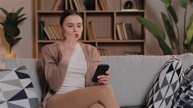 Sinirli, beyaz bir kadın kanepeye oturmuş elinde akıllı bir telefonla mesaj yazarken bağırıyor. Açık oturma odasında, arkada dekor olan bir evde çevrimiçi sohbet sırasında kızgınlığını ifade etmek..