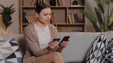 Elinde akıllı telefon ve kredi kartıyla kanepede oturan Kafkas bir kadın. Ödeme detaylarını çevrimiçi giriyorum. Huzurlu oturma odasında, arkasında raflar olan çevrimiçi alışveriş yapan odaklanmış bir kadın..