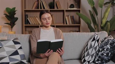 Huzurlu Kafkasyalı kadın kanepeye oturmuş, odaklanmış bir şekilde kitap okuyor. Sakin bir kadın sakin vakit geçirir ve evde rahat bir ortamda, arkasında raflar ve yeşil bitkilerle öğrenir. Boş hafta sonu.