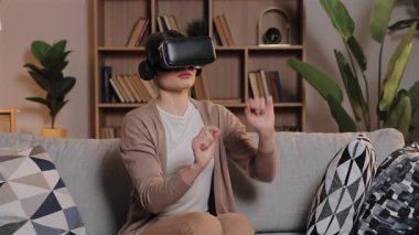 Sanal gerçeklik kulaklığı takan beyaz kadın evdeki kanepede oturup dijital alanla etkileşimde bulunuyor. Sanal objeyi raflarla dolu bir odada büyütmek için el kaldıran kadın..