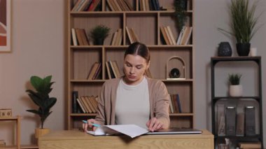 Ahşap masada oturan beyaz bir kadın gazete okuyor ve notlar alıyor. Ev işyerindeki belgelerin incelenmesi üzerine yoğunlaşan bir kadın dizüstü bilgisayarla ve kitaplarla dolu organize raflarla.